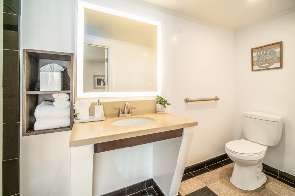 A modern bathroom with a lit vanity mirror, sink, shelf with towels, toilet, and wall art, featuring neutral tones and clean lines.