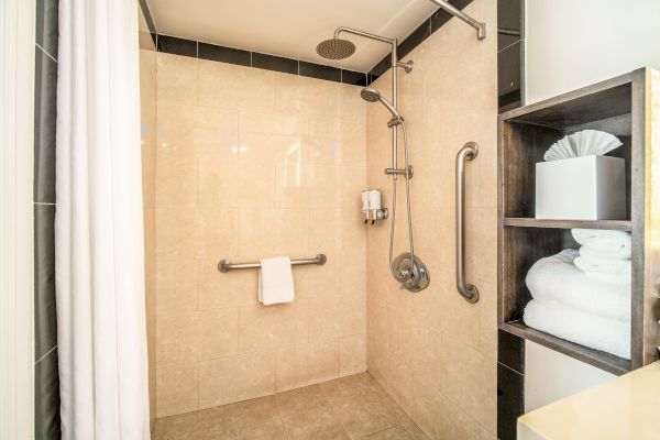 The image shows a modern shower with beige tiles, a wall-mounted showerhead, grab bars, and a towel. Nearby shelves hold folded towels and tissues.