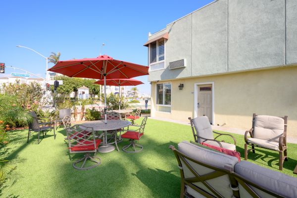 A courtyard with patio furniture, including chairs and tables under red umbrellas, located next to a two-story building on artificial grass.