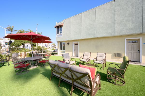 A patio with outdoor furniture, including cushioned chairs and a sofa, a table with a red umbrella, and artificial grass, near a building.