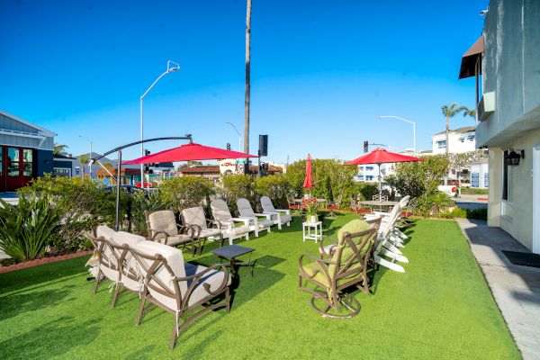 The image shows an outdoor seating area with chairs, umbrellas, artificial grass, and surrounding greenery under a clear blue sky.