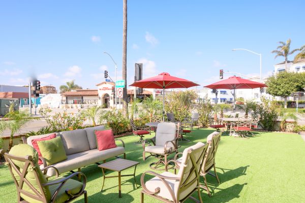 An outdoor seating area with chairs, tables, umbrellas, and lounges is set on a grass-like surface near a street with palm trees and buildings in the background.