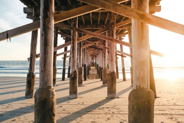 The image shows the view under a wooden pier on a beach during sunset, with long shadows cast by the pier's supports and the ocean visible in the distance.