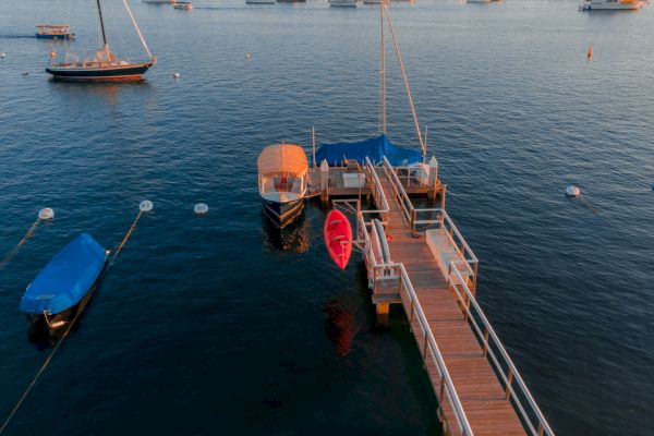 A wooden dock extends into calm waters with boats moored nearby, including a red kayak and others covered with blue tarps, in a serene setting.