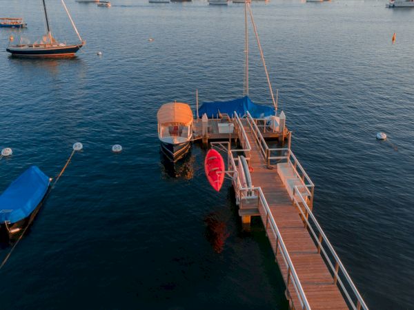 A wooden dock extends into calm waters with boats moored nearby, including a red kayak and others covered with blue tarps, in a serene setting.