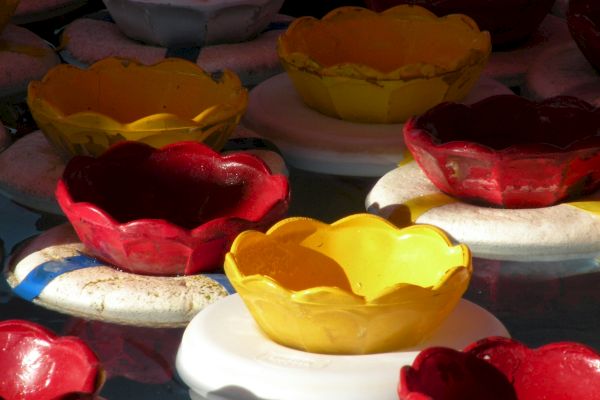 The image shows colorful floating flower-shaped bowls in red, yellow, and white, set on round white bases in a body of water, possibly for a festival.