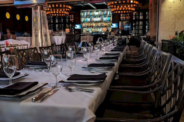 The image shows a long, elegantly set dining table with white tablecloths and black napkins in an outdoor restaurant setting, ready for guests.