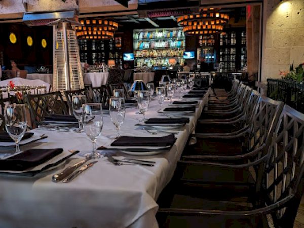The image shows a long, elegantly set dining table with white tablecloths and black napkins in an outdoor restaurant setting, ready for guests.