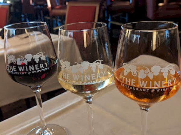Three wine glasses with different wines, labeled "The Winery," sit on a table in a restaurant with chairs and a person in the background.