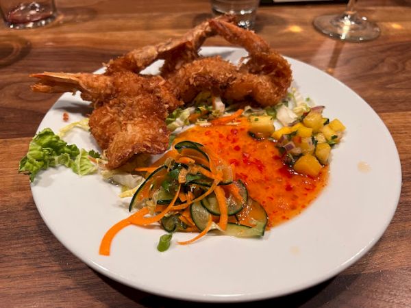 The image shows a plate with crispy fried shrimp, a tangy dipping sauce, a cucumber and carrot salad, and a side of mango salsa on a wooden table.