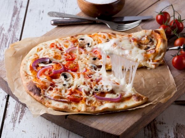 A pizza with melted cheese, tomatoes, onions, and mushrooms on a wooden board, with utensils and cherry tomatoes placed nearby, is shown.