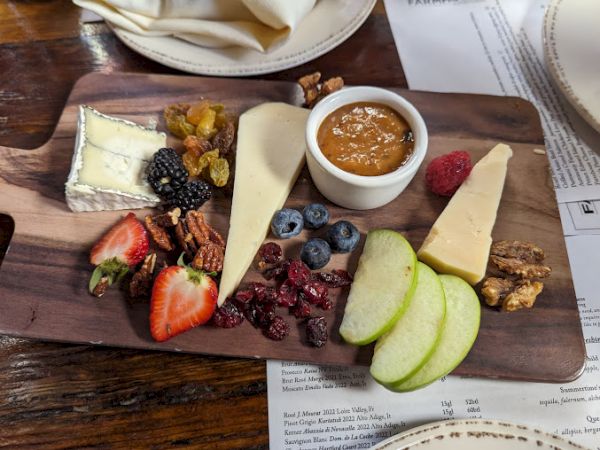 A wooden platter with cheeses, strawberries, blackberries, blueberries, green apple slices, various dried fruits, pecans, and a dipping sauce.