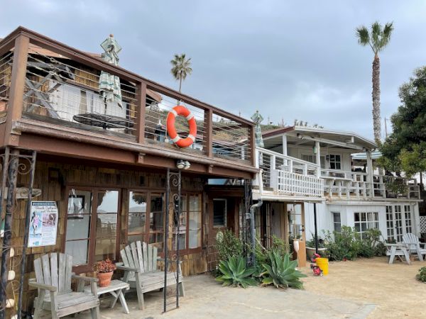 A coastal building with a lifebuoy, patio furniture, plants, and palm trees in the background under a cloudy sky.