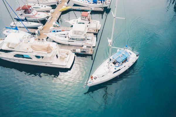 Aerial view of several boats docked at a marina, with one sailboat maneuvering away from the dock into the open water, creating ripples.