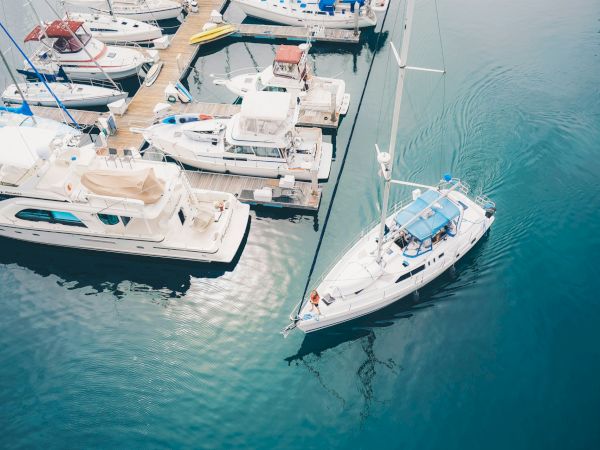 Aerial view of several boats docked at a marina, with one sailboat maneuvering away from the dock into the open water, creating ripples.