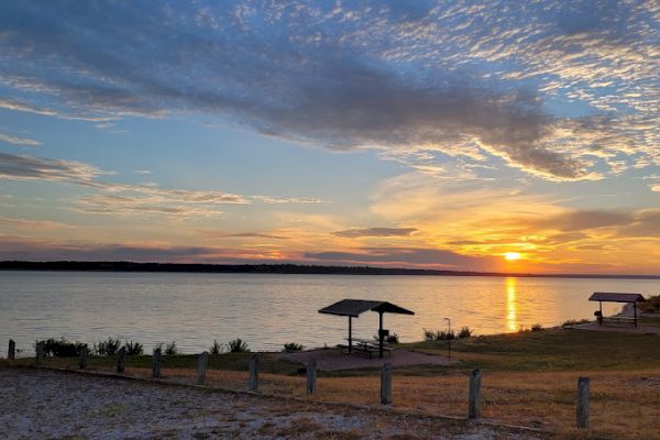 The image shows a serene lakeside sunset with wooden shelters, a tranquil body of water, and a partly cloudy sky with vibrant colors ending the sentence.