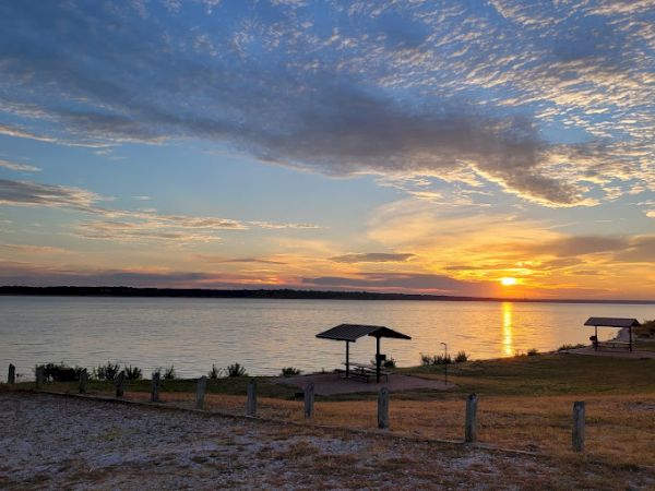 The image shows a serene lakeside sunset with wooden shelters, a tranquil body of water, and a partly cloudy sky with vibrant colors ending the sentence.