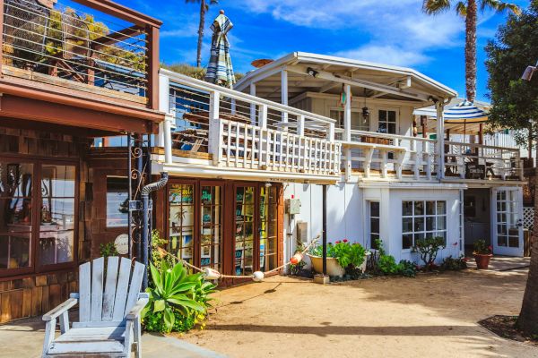 A two-story beach house with wooden accents, outdoor seating, lush plants, and a bright blue sky in the background.