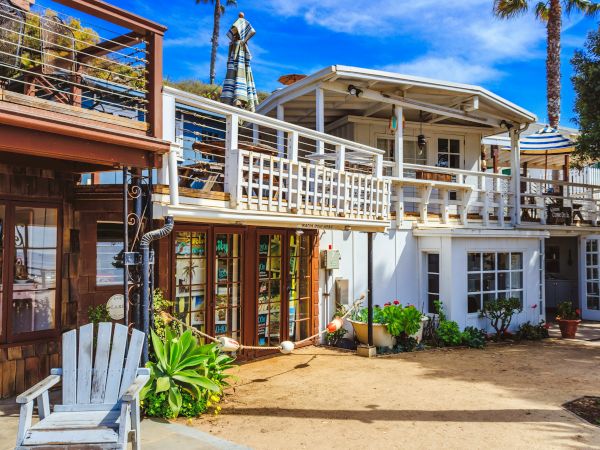 A two-story beach house with wooden accents, outdoor seating, lush plants, and a bright blue sky in the background.