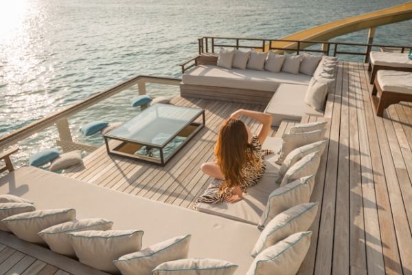 A woman sits in a luxurious outdoor seating area on a wooden deck overlooking a calm sea, with a glass table in the center and a slide leading into the water.