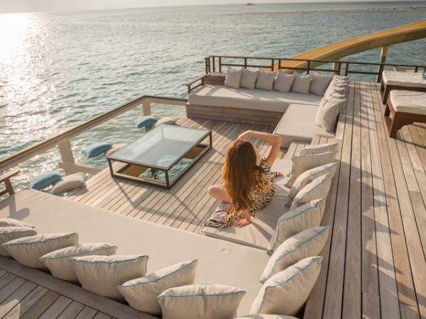 A woman sits in a luxurious outdoor seating area on a wooden deck overlooking a calm sea, with a glass table in the center and a slide leading into the water.