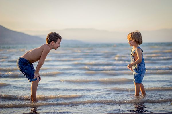 Two children are standing in shallow water at the beach, facing each other and wearing denim clothing, with a mountainous background.