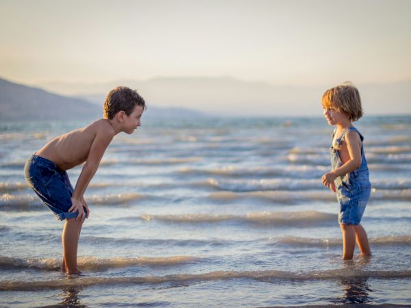 Two children are standing in shallow water at the beach, facing each other and wearing denim clothing, with a mountainous background.