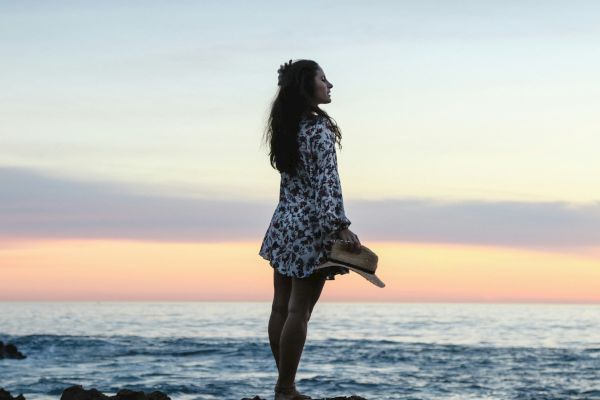 A woman in a floral dress holding a hat stands on a rocky shoreline, looking out at the ocean during sunset with colorful skies in the background.