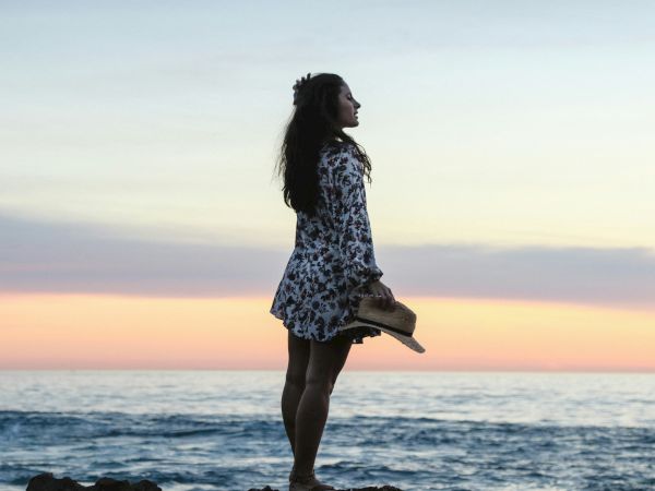A woman in a floral dress holding a hat stands on a rocky shoreline, looking out at the ocean during sunset with colorful skies in the background.