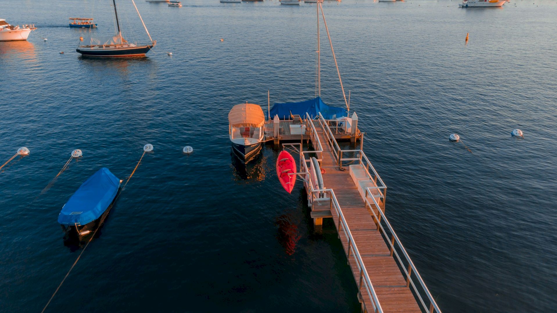 A dock with boats and a red kayak tied up on calm waters, with other boats anchored in the background, under a clear sky.