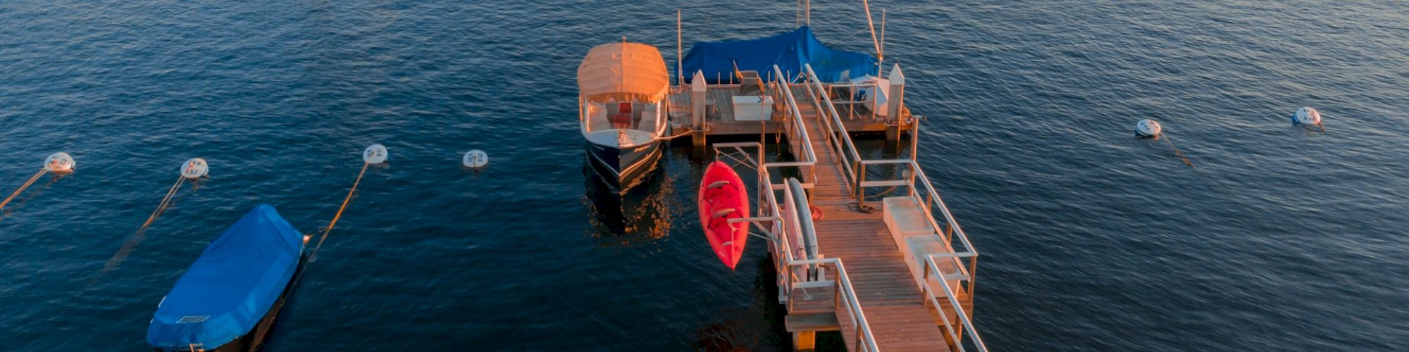 A dock with boats and a red kayak tied up on calm waters, with other boats anchored in the background, under a clear sky.