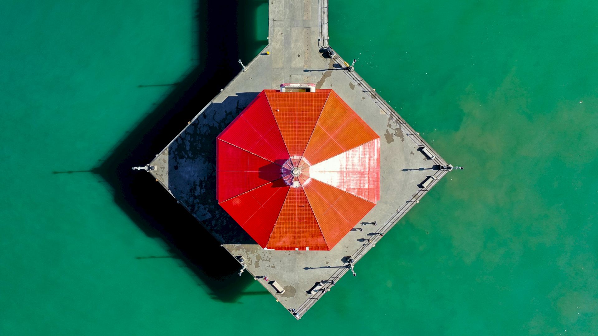 This image shows an aerial view of a red-roofed pavilion on a concrete pier surrounded by greenish-blue water.