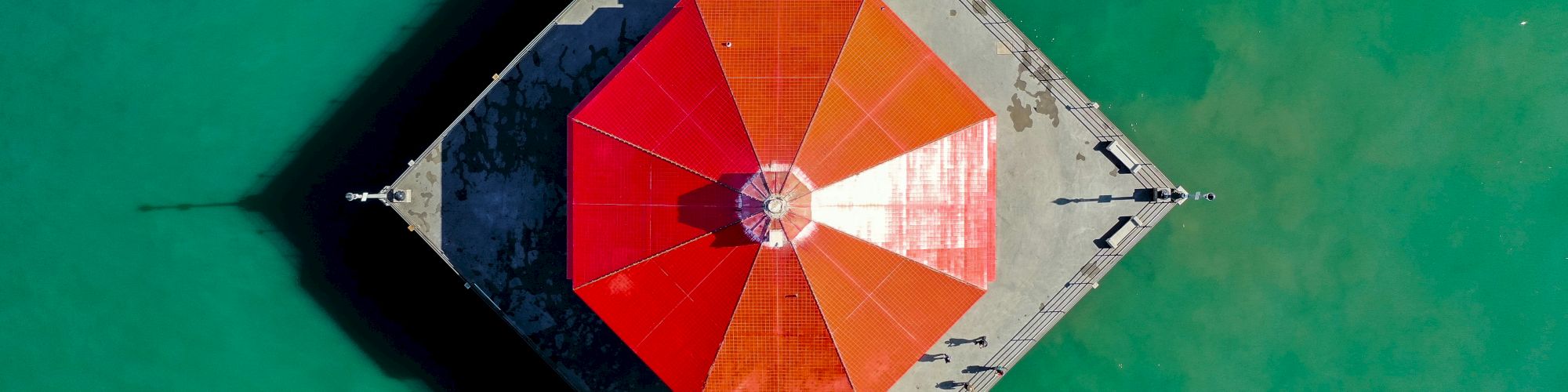 This image shows an aerial view of a red-roofed pavilion on a concrete pier surrounded by greenish-blue water.