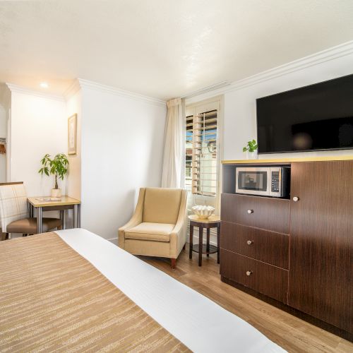 A hotel room with a bed, armchair, small dining table, TV above a dresser, and a microwave. The room has white walls and wood flooring.