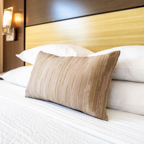 A neatly made bed with white sheets and pillows is topped with a decorative beige throw pillow; a modern headboard and wall light are visible.