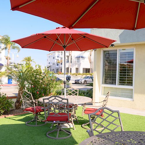 The image shows an outdoor seating area with red umbrellas, chairs, and tables on a patio with green artificial grass, near a building.