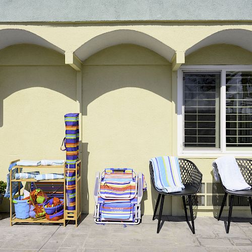 The image shows a patio with arched yellow walls. Outdoor chairs with towels, folded beach chairs, and a shelf with colorful beach supplies are visible.