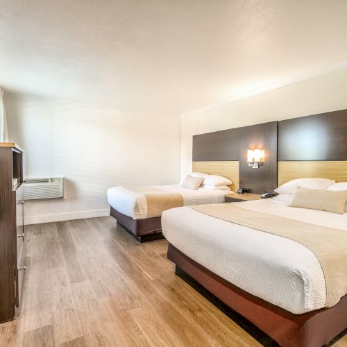 This image shows a hotel room with two double beds, a wall-mounted TV, an air conditioner, and wood flooring, featuring modern decor.