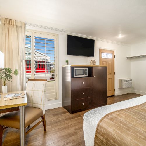 A cozy hotel room with a bed, a dresser with a microwave and TV, a table with a chair, and a window with curtains allowing natural light.