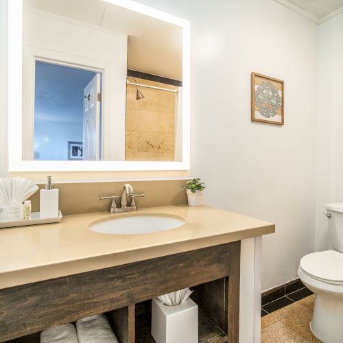 A modern bathroom with a lit mirror, sink, neatly arranged toiletries, toilet, and framed artwork on the wall, and an open doorway.