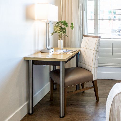 A small desk with a chair, lamp, plant, and reading materials is placed near a window with shutters in a bright room.