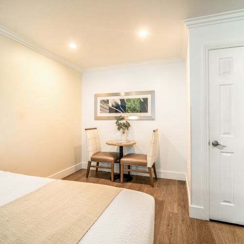 A cozy bedroom with a neatly made bed, a small table with two chairs, a door, and a picture on the wall above the table ends the sentence.