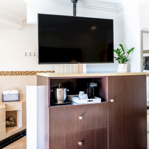 The image shows a hotel room with a mounted TV, a cabinet with a coffee maker and ice bucket, a jacuzzi tub, a mirror, and a small plant.