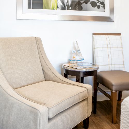 The image shows a beige armchair and a wooden chair with a cushioned seat, separated by a small round table holding water bottles and magazines.