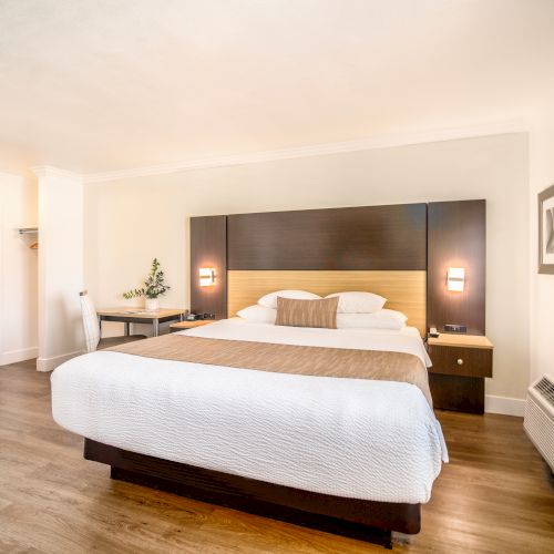 A modern hotel room with a large bed, two beside lamps, a small desk, a chair, and a wall-mounted TV, featuring wooden flooring and a clean design.