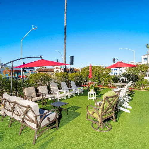 Outdoor seating area with chairs and loungers on artificial grass, shaded by red umbrellas, surrounded by buildings and palm trees, under a clear sky.