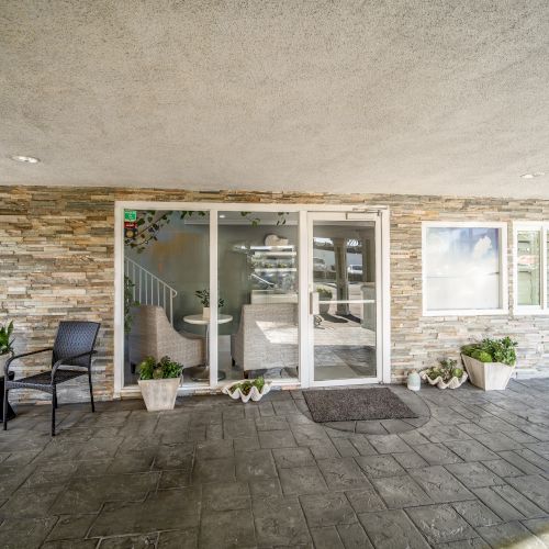 A covered entryway features glass double doors, stone walls, potted plants, a doormat, and a window, with two black chairs to the left.