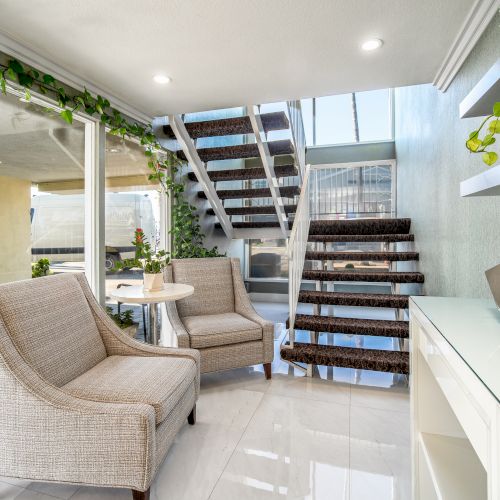 A modern, bright living space with two beige chairs, a small round table, a staircase, and various plants on shelves and surfaces, ending the sentence.