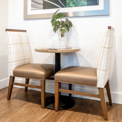 Two chairs sit around a small, round table with a potted plant, in a room with a wooden floor and an art piece on the wall.