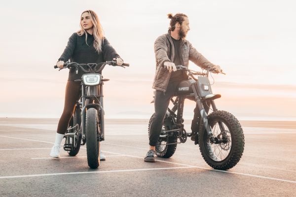 Two people are sitting on electric bikes in an open area, with a beautiful sunset in the background.